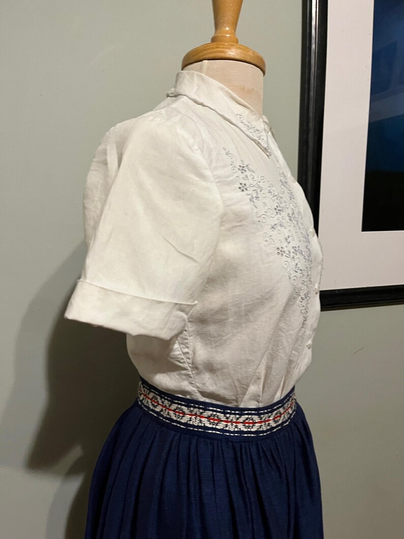 May include: A white short-sleeved blouse with embroidered floral details and a navy blue skirt with a white and red embroidered waistband.