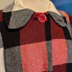 May include: A vintage coat with a large red, black, and gray plaid pattern. The coat features a cream-colored collar and a red, square-shaped button. The fabric appears to be a woven material.
