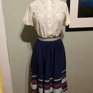 May include: A white embroidered blouse with a collar and short sleeves paired with a navy blue skirt with white and red embroidered trim.