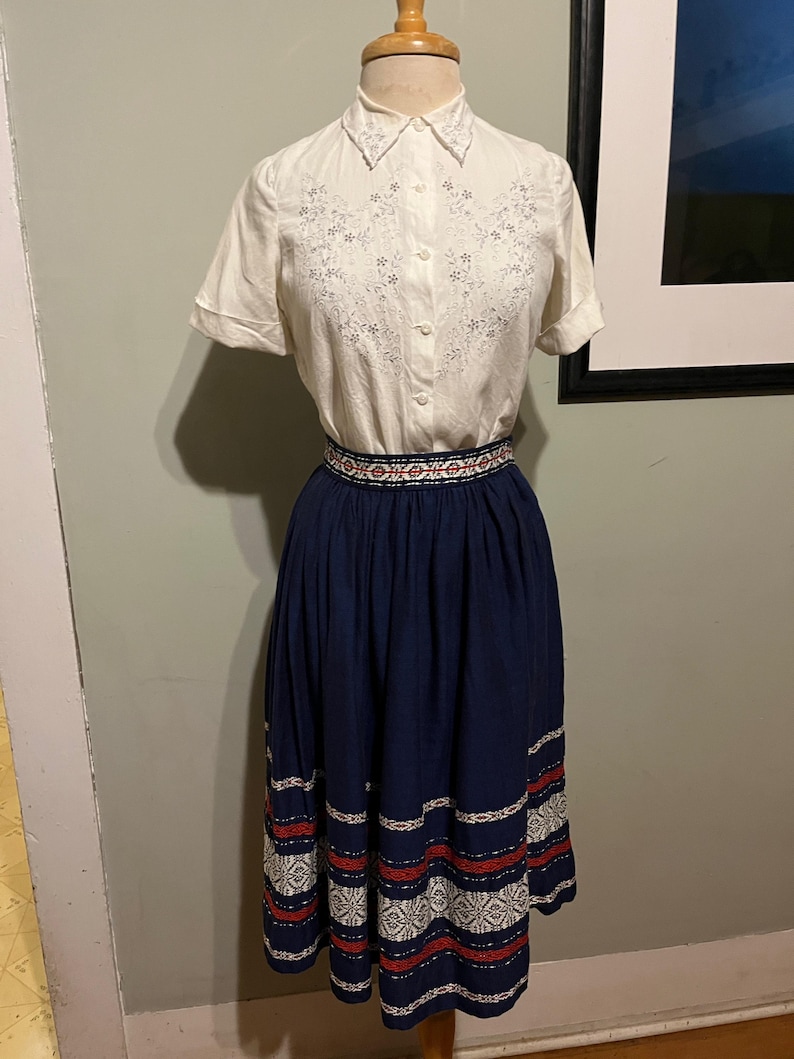 May include: A white embroidered blouse with a collar and short sleeves paired with a navy blue skirt with white and red embroidered trim.