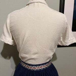 May include: A white short-sleeved button-up shirt with a collar. The shirt is worn over a navy blue skirt with a white and red embroidered band around the waist.
