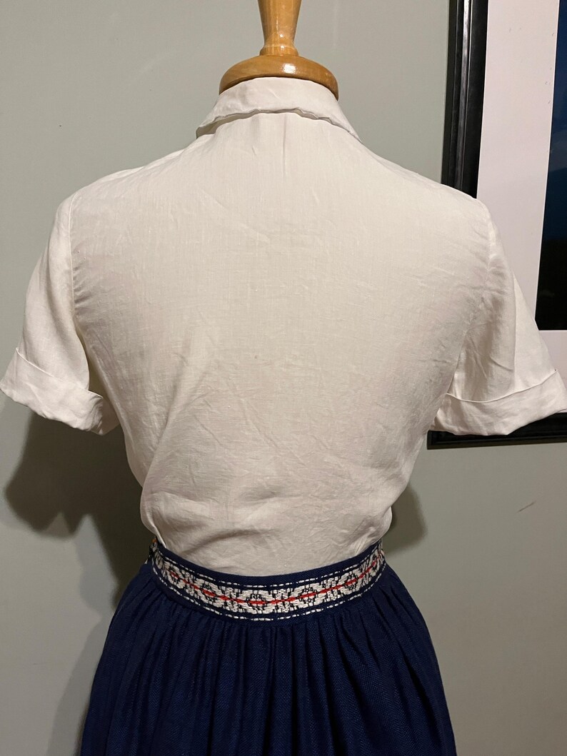May include: A white short-sleeved button-up shirt with a collar. The shirt is worn over a navy blue skirt with a white and red embroidered band around the waist.