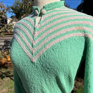 Puede incluir: Un suéter verde vintage tejido con un cuello alto y un diseño en forma de V en beige claro. El suéter tiene mangas largas y un cinturón burdeos. El cuello presenta dos pequeños botones redondos. El suéter se exhibe en un maniquí.