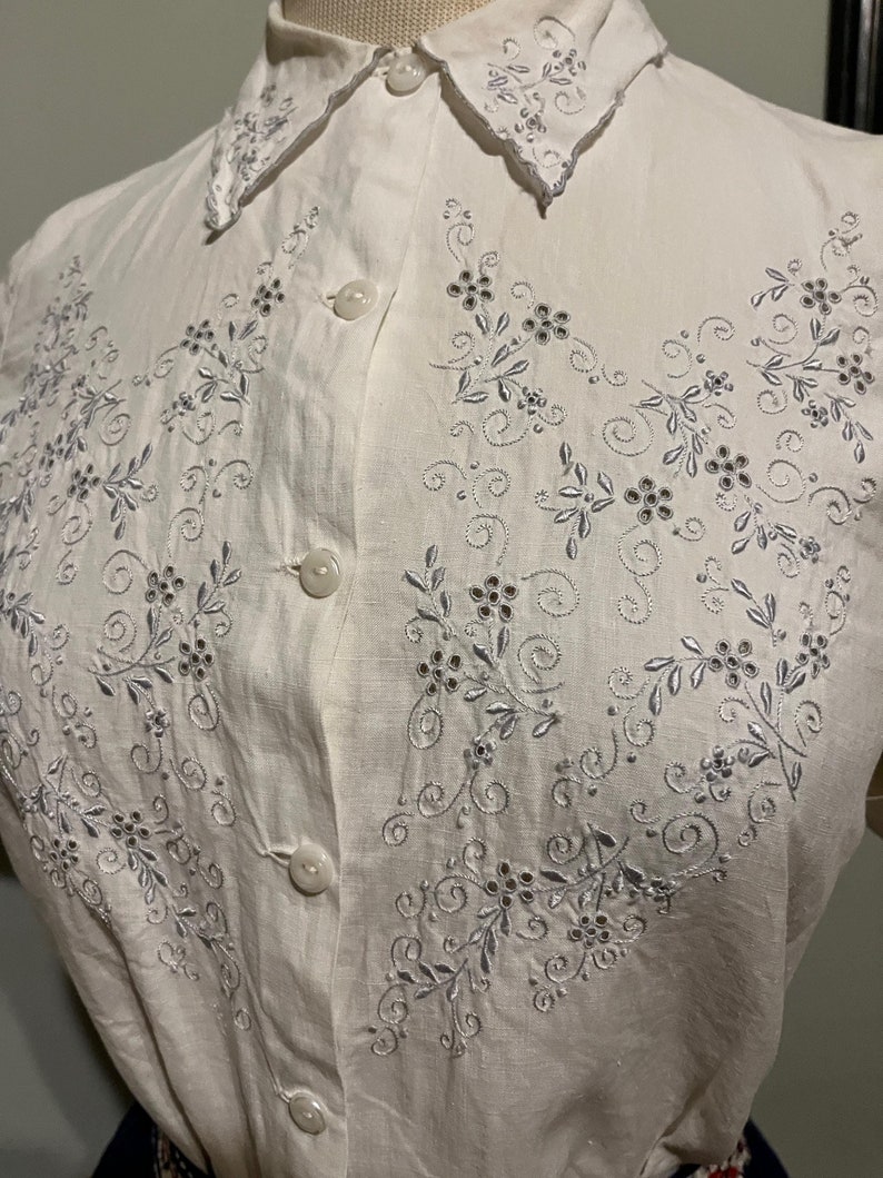May include: A white linen button-down shirt with a delicate floral embroidery design in silver thread. The embroidery features small flowers and swirls, creating a vintage and elegant look.