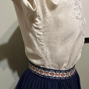 May include: A white blouse with embroidered floral details and a navy blue skirt with a red and white patterned waistband.