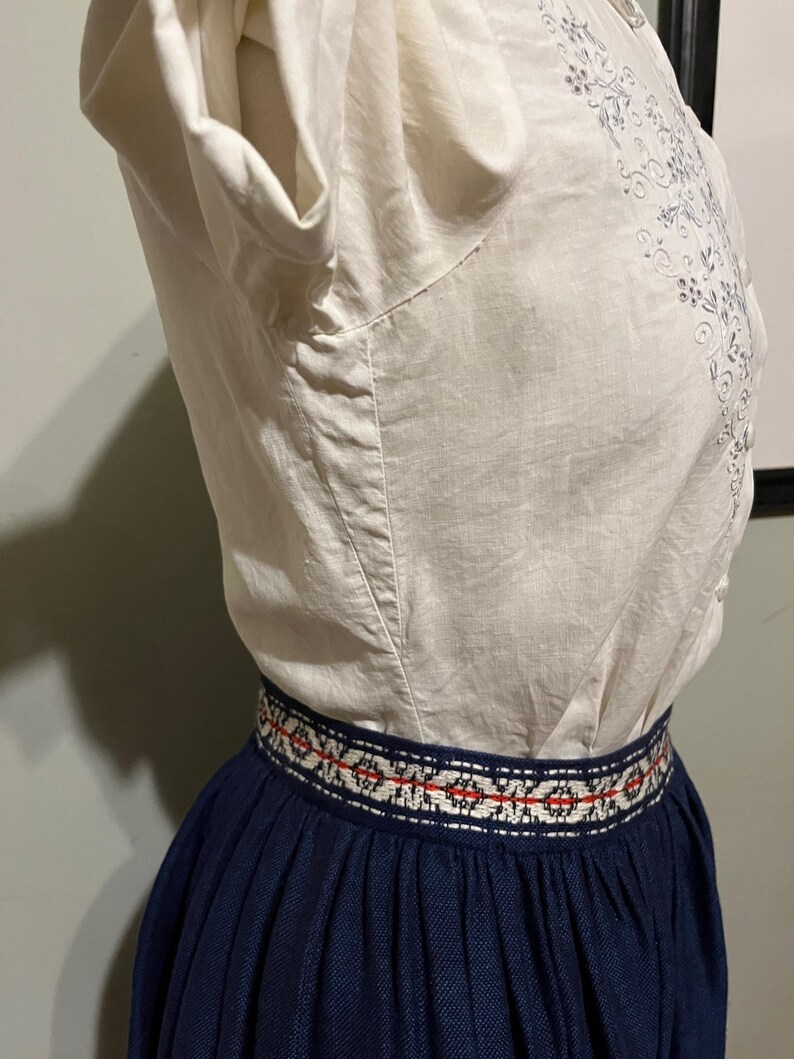 May include: A white blouse with embroidered floral details and a navy blue skirt with a red and white patterned waistband.