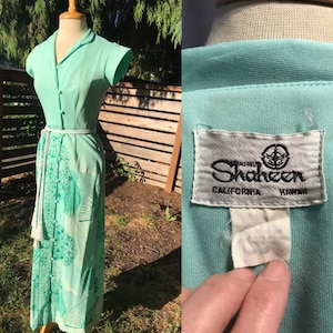 May include: A mint green maxi dress with a white rope belt and a green floral print. The dress has short sleeves and a button-up front. The label reads "Alfred Shaheen California Hawaii".