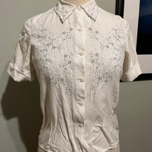 May include: A white button-up shirt with short sleeves and a delicate floral embroidery design in silver thread. The shirt has a pointed collar and a fitted silhouette.