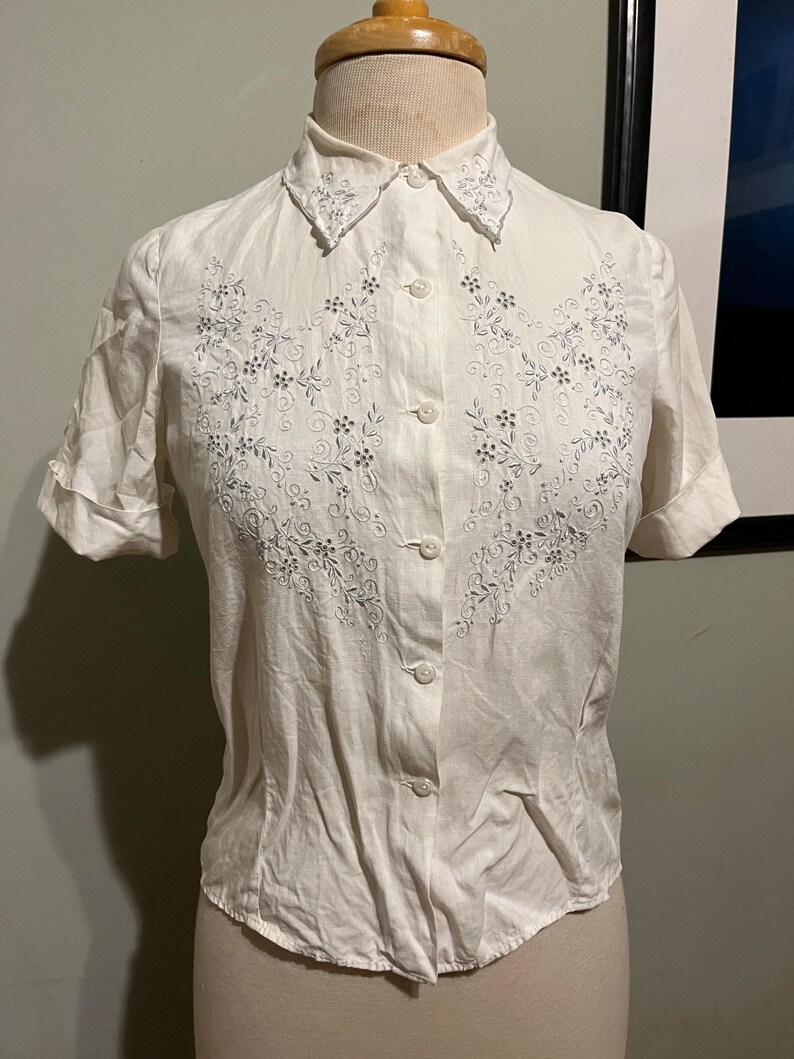 May include: A white button-up shirt with short sleeves and a delicate floral embroidery design in silver thread. The shirt has a pointed collar and a fitted silhouette.