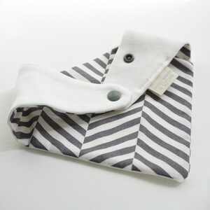 May include: A white and gray striped bib with a white cotton lining and two snap closures. The bib has a label that reads "GOTS Certified Made in Scotland".