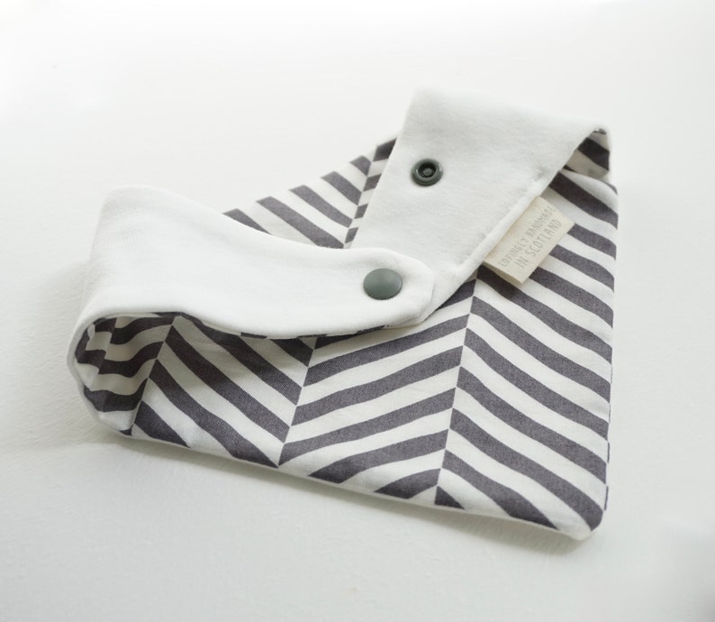 May include: A white and gray striped bib with a white cotton lining and two snap closures. The bib has a label that reads "GOTS Certified Made in Scotland".