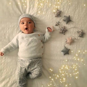 May include: A baby wearing a light gray knit sweater with a star design, matching pants with a star pattern, and a gray beanie. The baby is lying on a white surface, with a star garland and decorative sparkles.