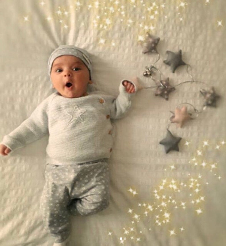 May include: A baby wearing a light gray knit sweater with a star design, matching pants with a star pattern, and a gray beanie. The baby is lying on a white surface, with a star garland and decorative sparkles.