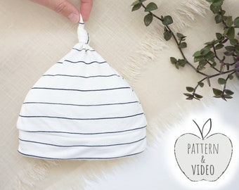 Puede incluir: Un gorro de punto blanco y negro a rayas con un nudo en la parte superior. El gorro está sobre una superficie blanca con una planta verde en el fondo. El texto "PATTERN & VIDEO" está sobre una forma de manzana blanca.