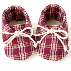 May include: A pair of burgundy and cream plaid baby booties with white laces tied in a bow.