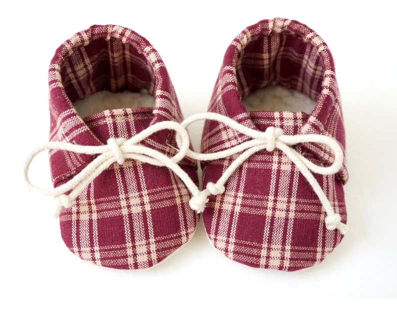 May include: A pair of burgundy and cream plaid baby booties with white laces tied in a bow.