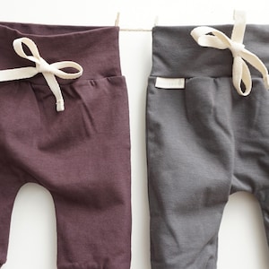 May include: Two pairs of baby pants in plum and charcoal gray, hanging on a clothesline. Each pair has a drawstring waist with a cream-colored tie and ribbed cuffs. The pants are made of soft fabric.