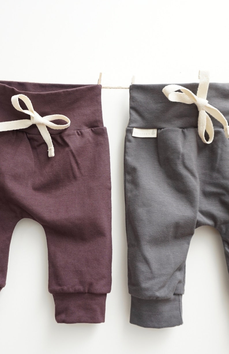 May include: Two pairs of baby pants in plum and charcoal gray, hanging on a clothesline. Each pair has a drawstring waist with a cream-colored tie and ribbed cuffs. The pants are made of soft fabric.