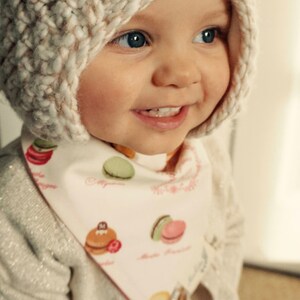 May include: A baby wearing a white knitted hat with a brown and white pattern and a white bib with a colorful macaron pattern. The bib has the text "Macarons" and "Paris" on it.