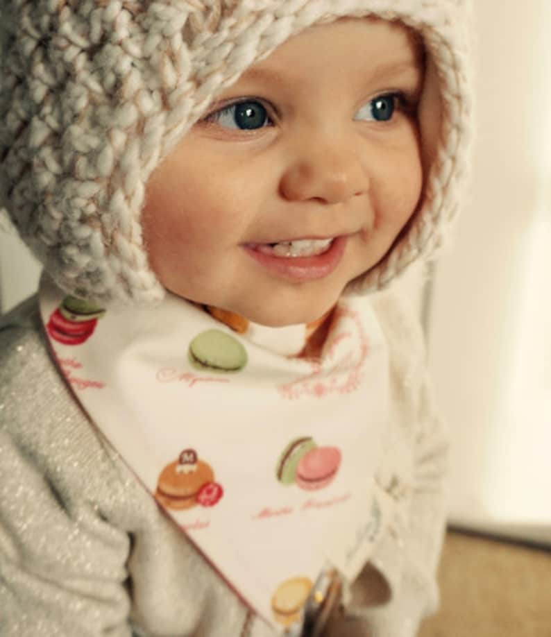 May include: A baby wearing a white knitted hat with a brown and white pattern and a white bib with a colorful macaron pattern. The bib has the text "Macarons" and "Paris" on it.