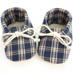 May include: A pair of baby booties with a plaid pattern in navy blue and beige. The booties have a white lace-up closure.
