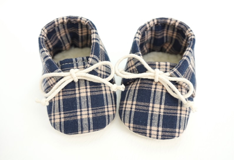 May include: A pair of baby booties with a plaid pattern in navy blue and beige. The booties have a white lace-up closure.