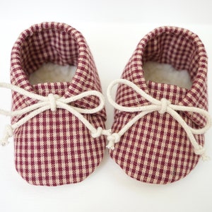 May include: A pair of baby moccasins with a burgundy and cream gingham pattern and white laces tied in a bow.