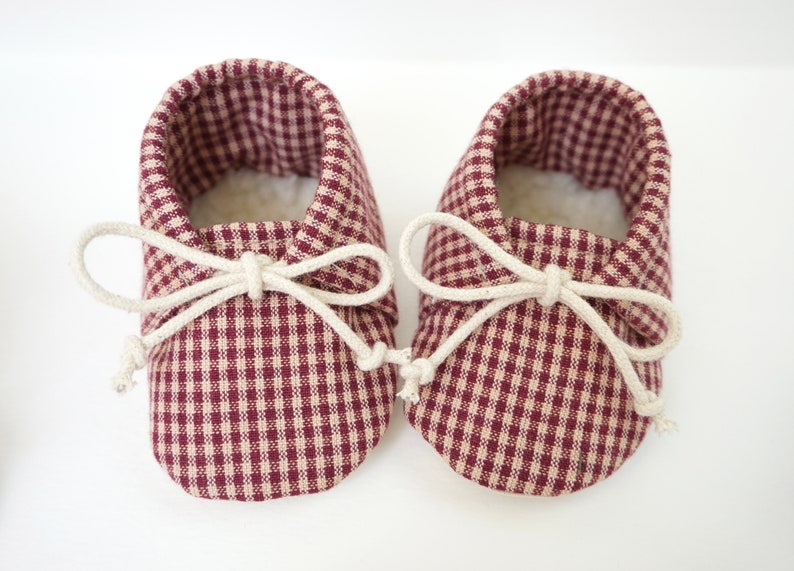 May include: A pair of baby moccasins with a burgundy and cream gingham pattern and white laces tied in a bow.