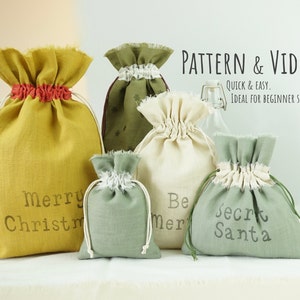 Puede incluir: Cinco bolsas de cordón hechas de tela de lino natural con diferentes colores y diseños. Las bolsas están decoradas con el texto "Merry Christmas", "Be Merry" y "Secret Santa".