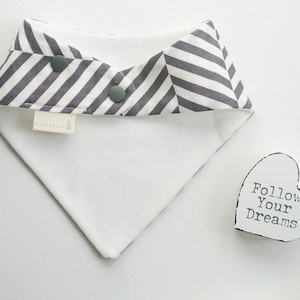 May include: A white and gray striped bandana bib with two snap closures. The bib has a white label with the word "Noah" printed on it. The bib is lying on a white surface next to a white heart-shaped sign with the text "Follow Your Dreams" printed on it.