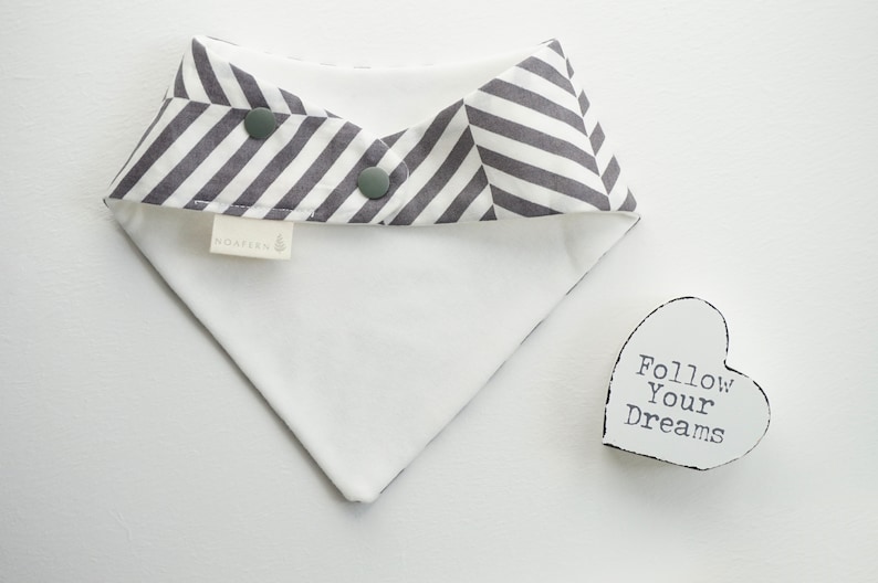 May include: A white and gray striped bandana bib with two snap closures. The bib has a white label with the word "Noah" printed on it. The bib is lying on a white surface next to a white heart-shaped sign with the text "Follow Your Dreams" printed on it.