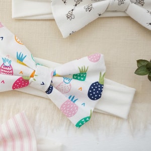 Puede incluir: Una diadema blanca con un lazo hecho de tela con estampado de pi&ntilde;a. El lazo est&aacute; atado en un nudo y la tela tiene una variedad de colores, incluyendo rosa, verde, azul, amarillo y naranja.