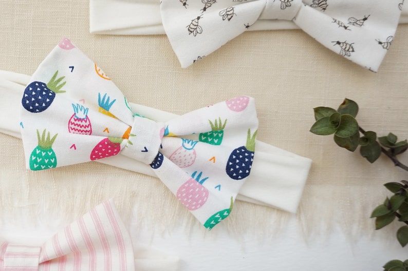 Puede incluir: Una diadema blanca con un lazo hecho de tela con estampado de pi&ntilde;a. El lazo est&aacute; atado en un nudo y la tela tiene una variedad de colores, incluyendo rosa, verde, azul, amarillo y naranja.