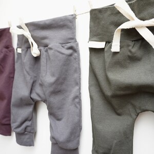 May include: Three pairs of baby pants in shades of purple, gray, and olive green hang from a clothesline. Each pair has a wide waistband with a white drawstring and elastic cuffs at the ankles. The pants are made of soft fabric.