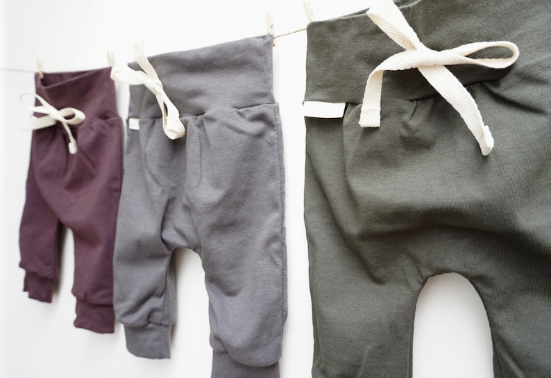 May include: Three pairs of baby pants in shades of purple, gray, and olive green hang from a clothesline. Each pair has a wide waistband with a white drawstring and elastic cuffs at the ankles. The pants are made of soft fabric.