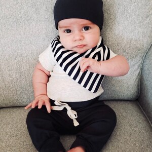 May include: A baby wearing a black beanie, a white and black striped bib, a grey short-sleeved shirt, and black pants.
