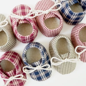 May include: Eight pairs of baby booties in various plaid patterns, including red and white, blue and white, and green and white. The booties have white laces and are made of soft fabric.