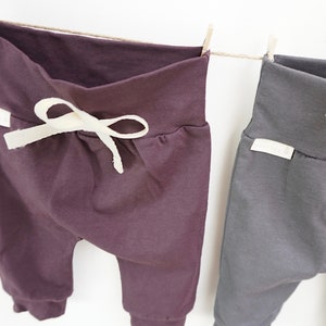 May include: Two pairs of baby pants hanging on a clothesline. One pair is a deep purple with a cream-colored drawstring, and the other is a dark gray. Both have a folded waistband and cuffed ankles.