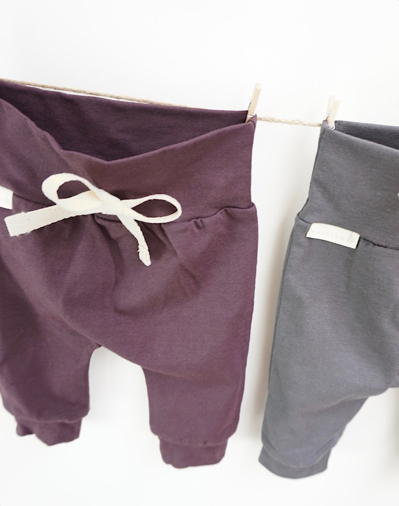 May include: Two pairs of baby pants hanging on a clothesline. One pair is a deep purple with a cream-colored drawstring, and the other is a dark gray. Both have a folded waistband and cuffed ankles.