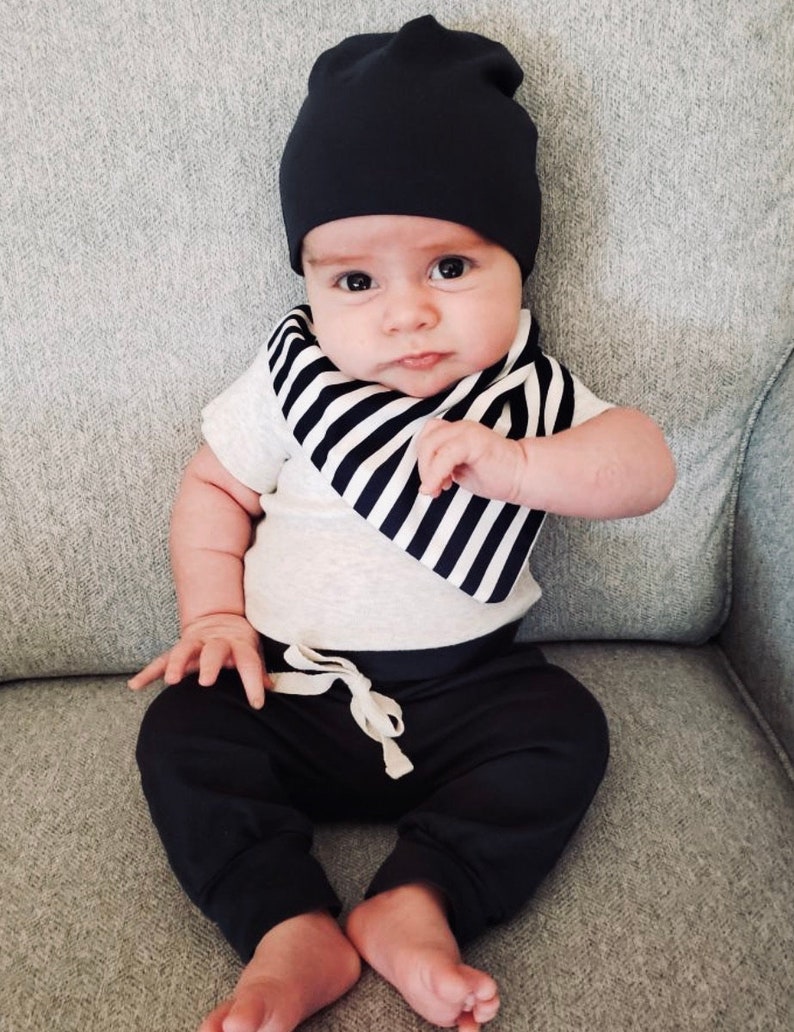 May include: A baby wearing a black beanie, a white t-shirt with a black and white striped bib, and black sweatpants with a drawstring. The baby is sitting on a gray couch.
