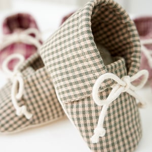 May include: A pair of baby moccasins in a green and white gingham pattern with white laces tied in a bow. The moccasins are soft and comfortable for little feet.