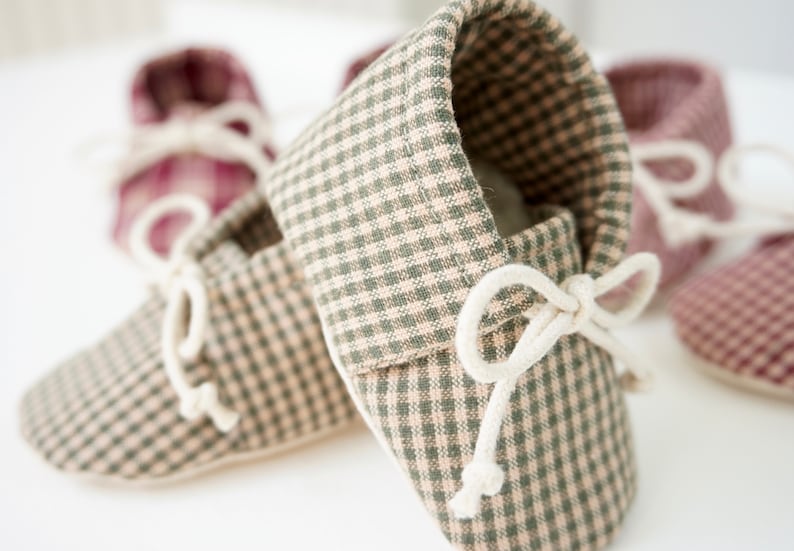 May include: A pair of baby moccasins in a green and white gingham pattern with white laces tied in a bow. The moccasins are soft and comfortable for little feet.