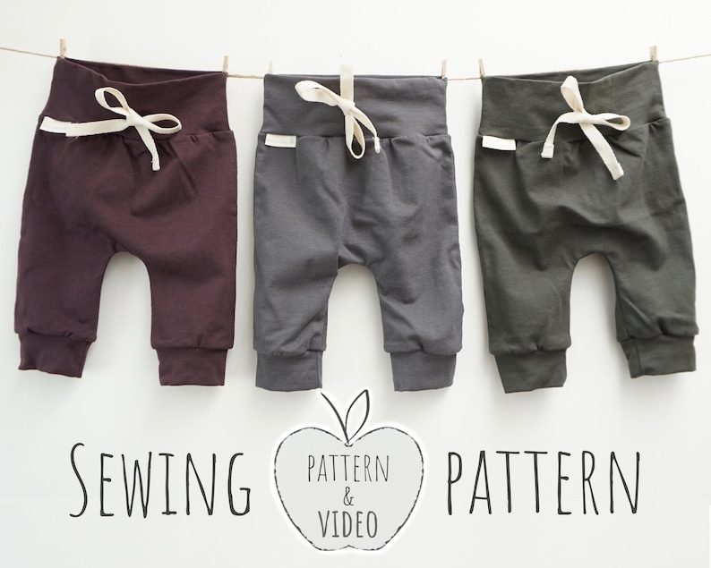 May include: Three pairs of baby pants in burgundy, gray, and olive green hang from a clothesline. Each pair has a high waistband with a drawstring and a small white tag. The text "SEWING PATTERN & VIDEO PATTERN" is below the pants.