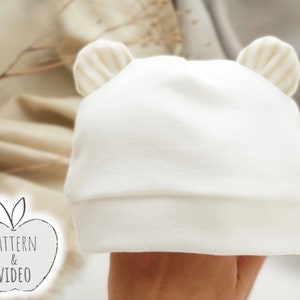 Può includere: Cappellino bianco per neonato con alette beige. Il cappello è realizzato in tessuto morbido ed elastico e ha un design semplice.