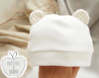 May include: White baby hat with beige ear flaps. The hat is made of soft, stretchy fabric and has a simple design.