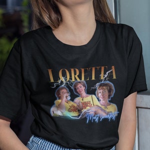 May include: A black t-shirt featuring a graphic print of three women wearing yellow shirts. The text "LORETTA" is printed in gold above the image. The text "Most swankiest" is printed in blue below the image.
