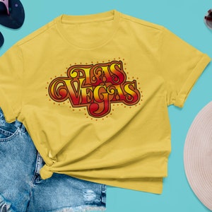 May include: Yellow t-shirt with the text "Las Vegas" in a retro, stylized font. The text is in shades of red and orange with a gold outline. The shirt is paired with denim shorts, sunglasses, and sandals.