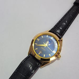 Peut inclure: Une montre-bracelet dorée avec un cadran bleu et un bracelet en cuir noir. Le cadran de la montre comporte des chiffres romains et le mot "AMERICA". Le bracelet est texturé. La montre est inclinée.