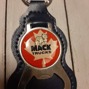 Op de afbeelding: Een blauwe leren sleutelhanger met een zilveren flesopener en een rood, wit en gouden rond embleem met een bulldog, een esdoornblad en de tekst "MACK TRUCKS".