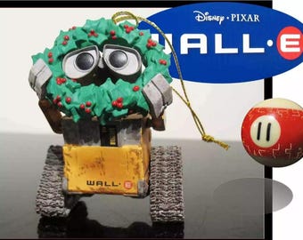 Może przedstawiać: Choinkowa ozdoba przedstawiająca WALL-E, robota z filmu Disneya Pixara WALL-E. Robot nosi zielony wieniec z ostrokrzewu z czerwonymi jagodami. Ozdoba jest wykonana z plastiku i ma złotą sznurkę do zawieszenia.